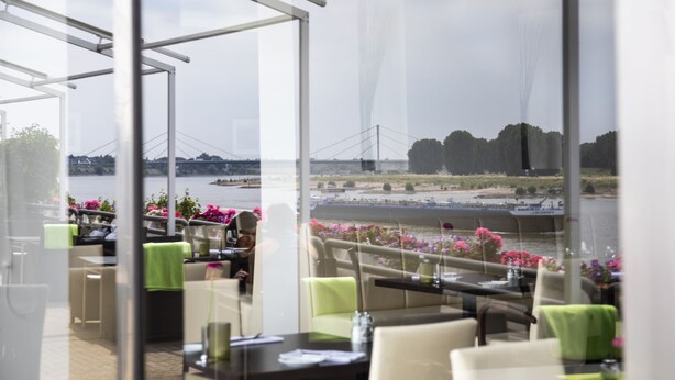 A restaurant with outdoor seating overlooking a river, featuring a glass wall for views. - Hotel Rheingarten Duisburg (Duisburg)