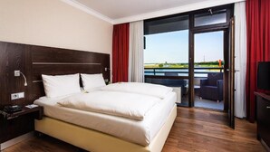 A hotel room with a large bed, a nightstand, a TV, and a balcony with a view of the water. - Hotel Rheingarten Duisburg (Duisburg)