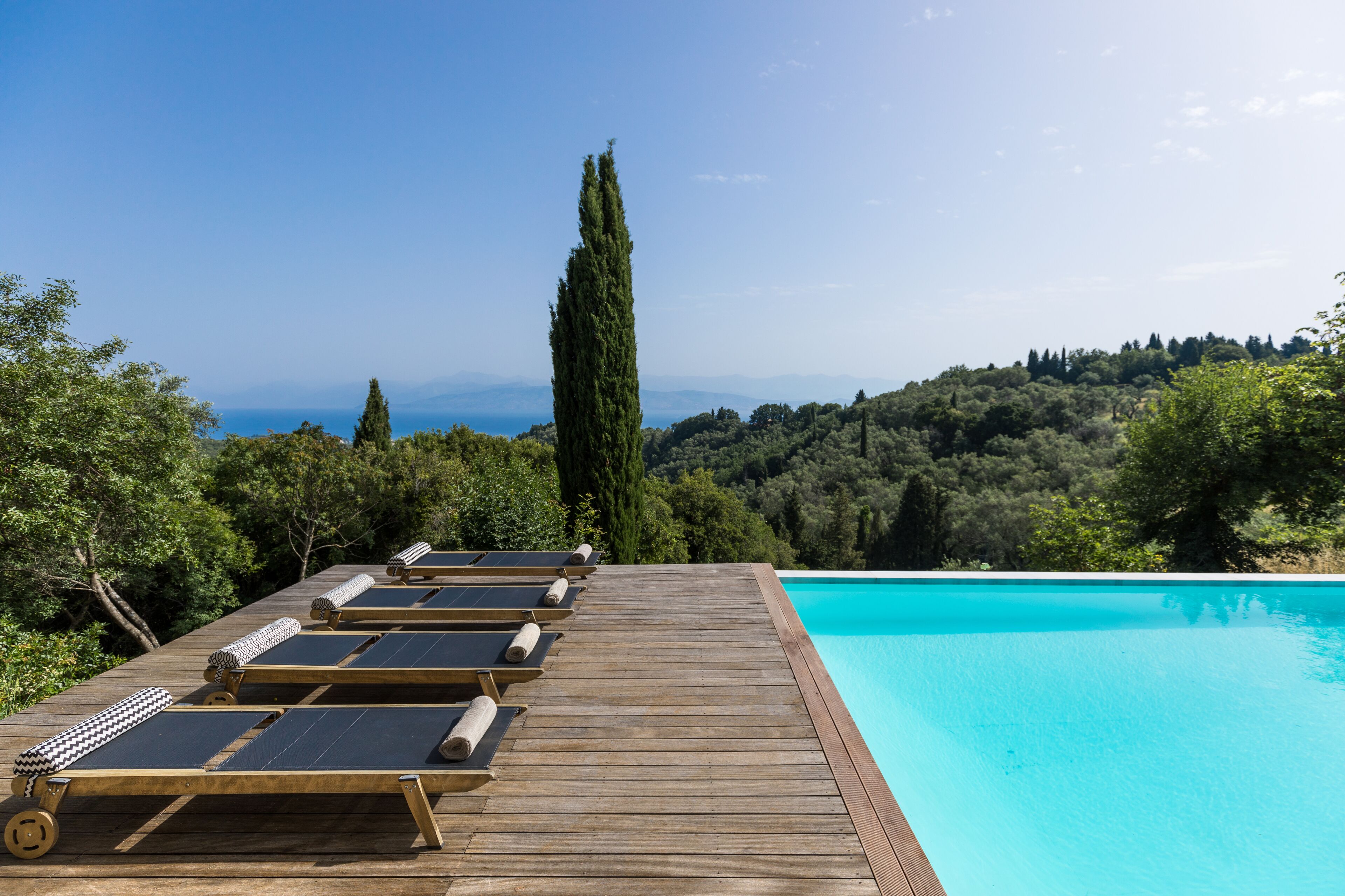 Terraza de madera con tumbonas que da a una piscina azul cristalina y una vista panorámica de una ladera boscosa.