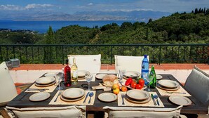 Ein Esszimmertisch draußen mit Geschirr, Besteck und einer Flasche, mit Blick auf eine schöne Landschaft mit Bäumen und Meer.