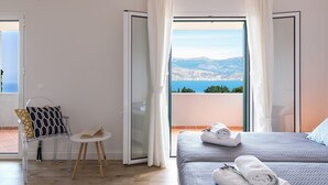 Ein Hotelzimmer mit einem Bett, einem kleinen Tisch und einem Stuhl. Durch das Fenster ist Meer und Berge zu sehen.