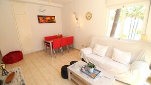 Ein Wohnzimmer mit weißem Sofa, einem roten Esstisch, einem Couchtisch und einem Fenster mit Blick auf Bäume.