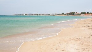 Ein Strand mit klarem Wasser, sandigem Ufer und Gebäuden im Hintergrund.