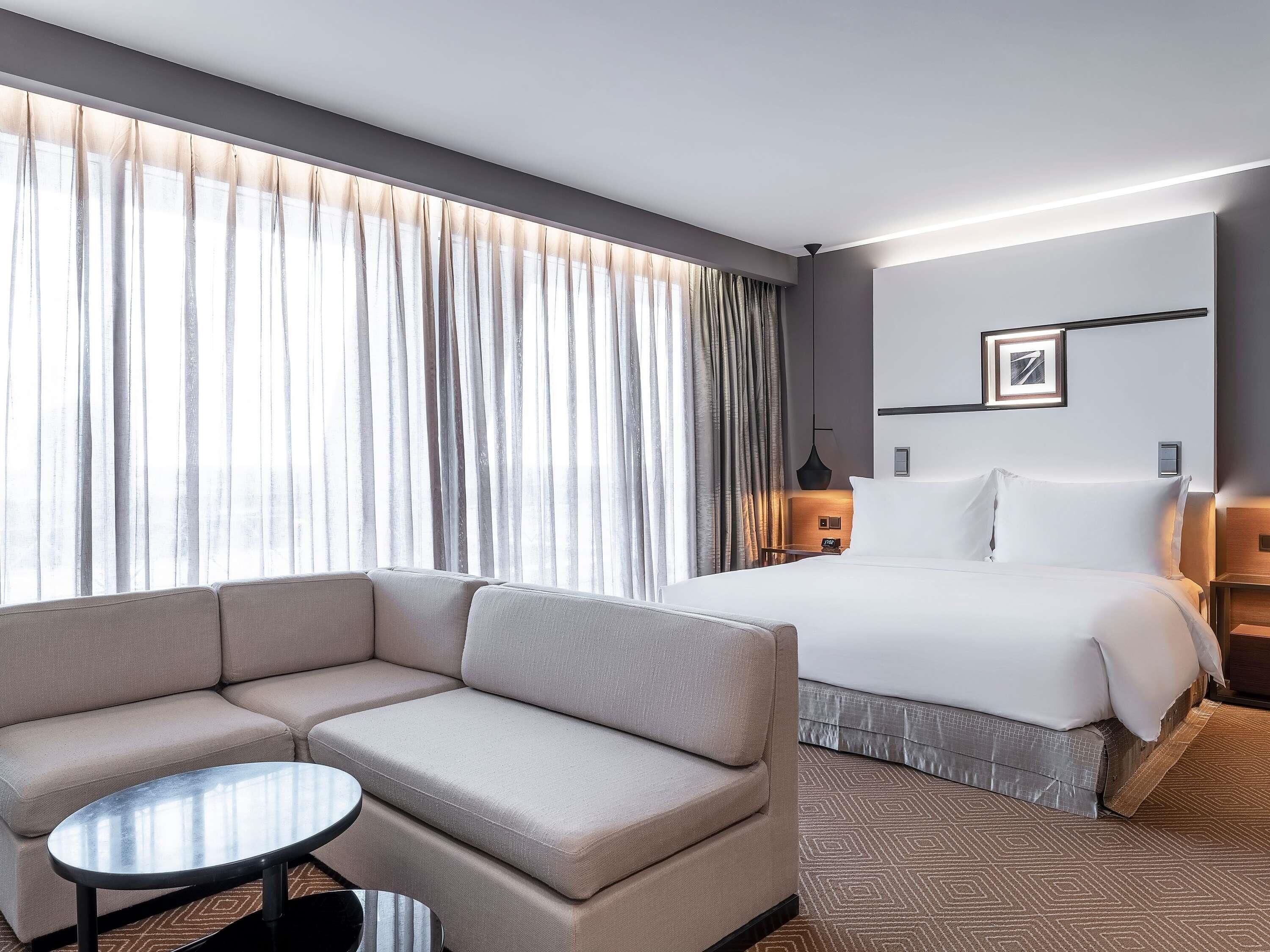 A hotel room with a bed, a sofa, a small table, and a large window with curtains.
