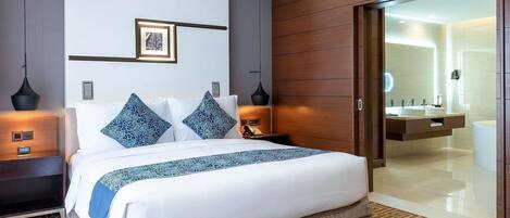 A modern hotel room with a large bed, bedside tables, a nightstand, a wall-mounted picture, and a bathroom with a large mirror and sink.