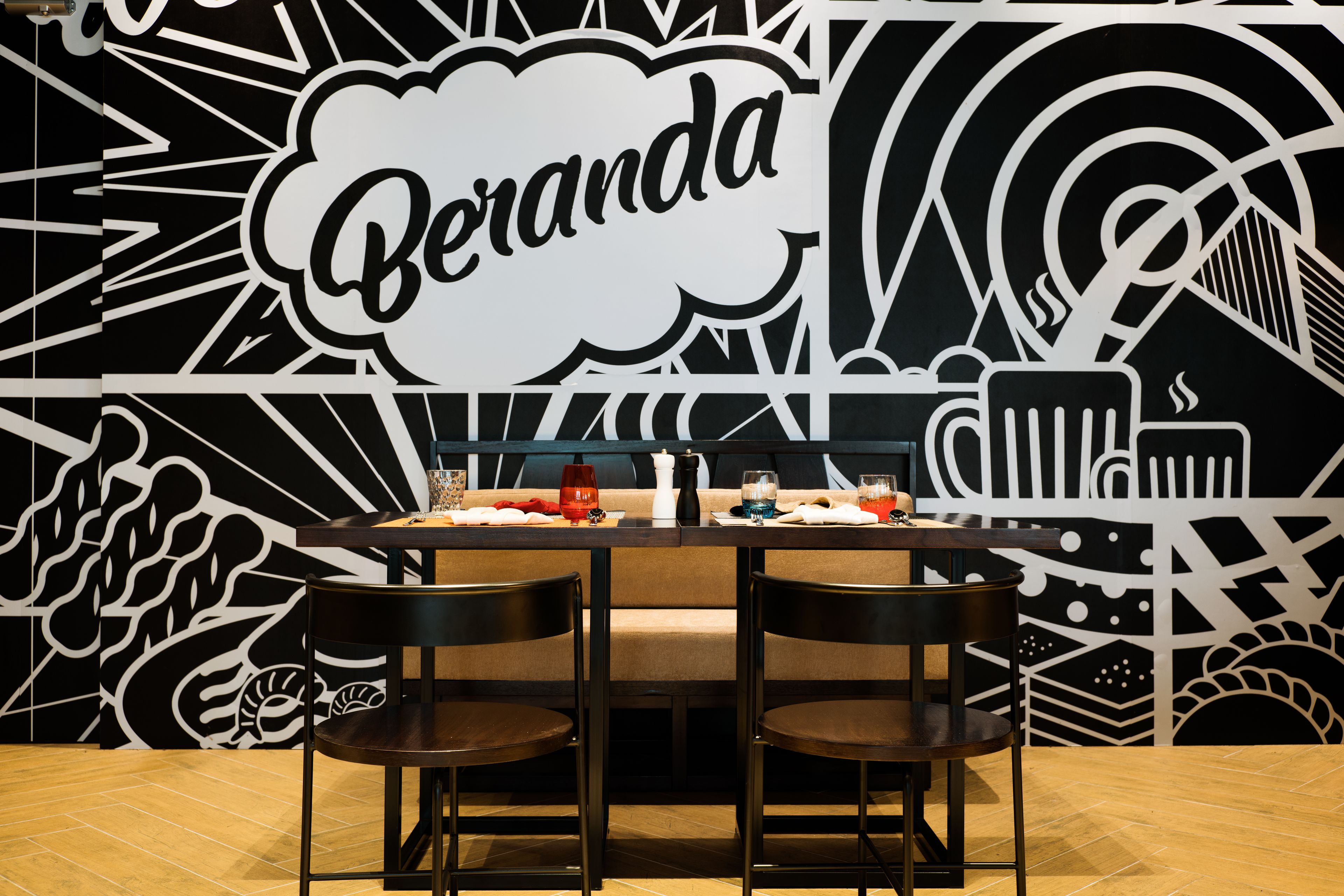 A dining area with a table set for two, a black and white mural with the word "Beranda" prominently displayed, and a patterned floor.