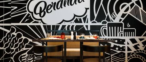 A dining area with a table set for two, a black and white mural with the word "Beranda" prominently displayed, and a patterned floor.