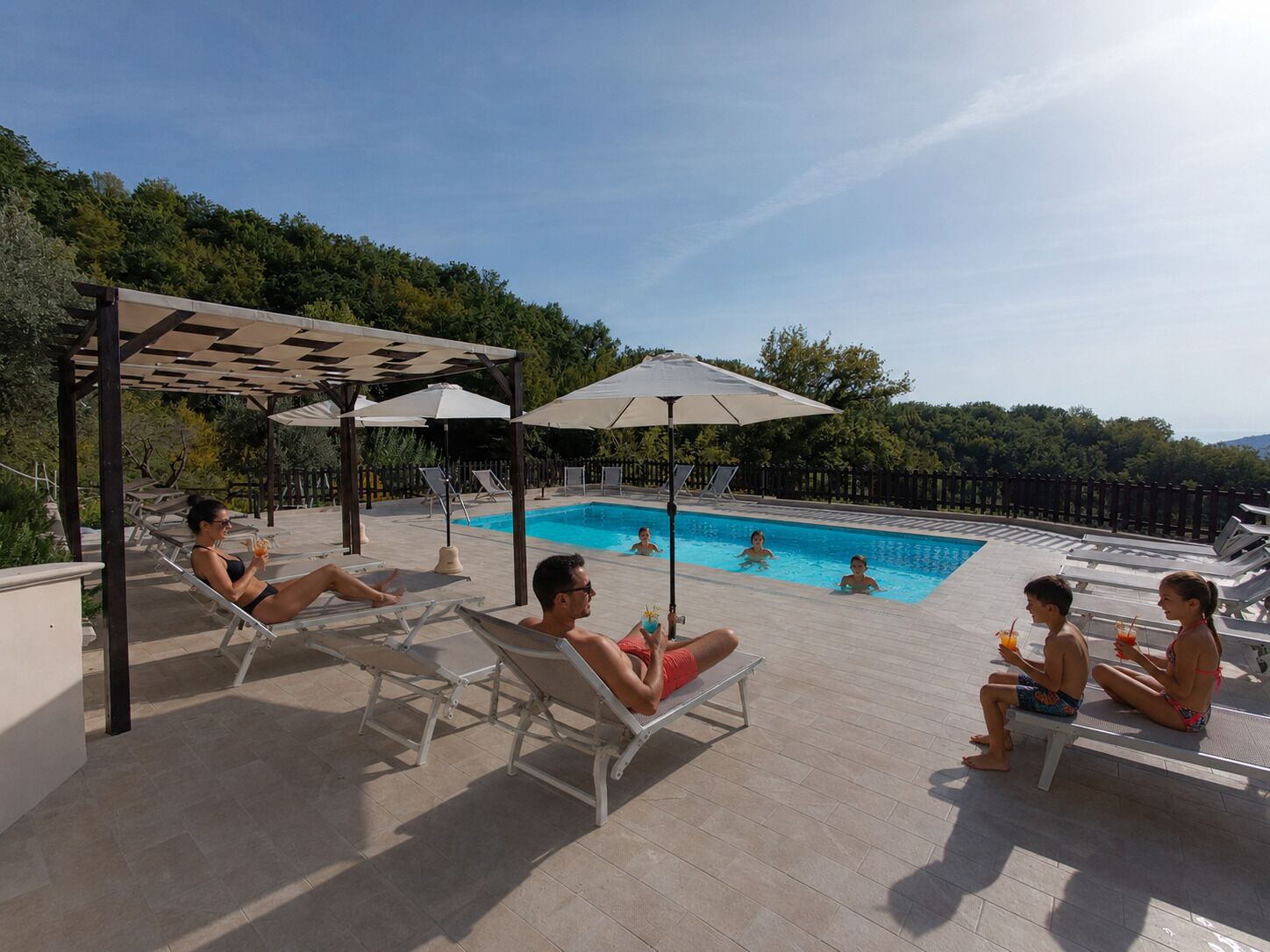 Area piscina con lettini e ombrelloni, circondata da un pontile in legno e alberi.