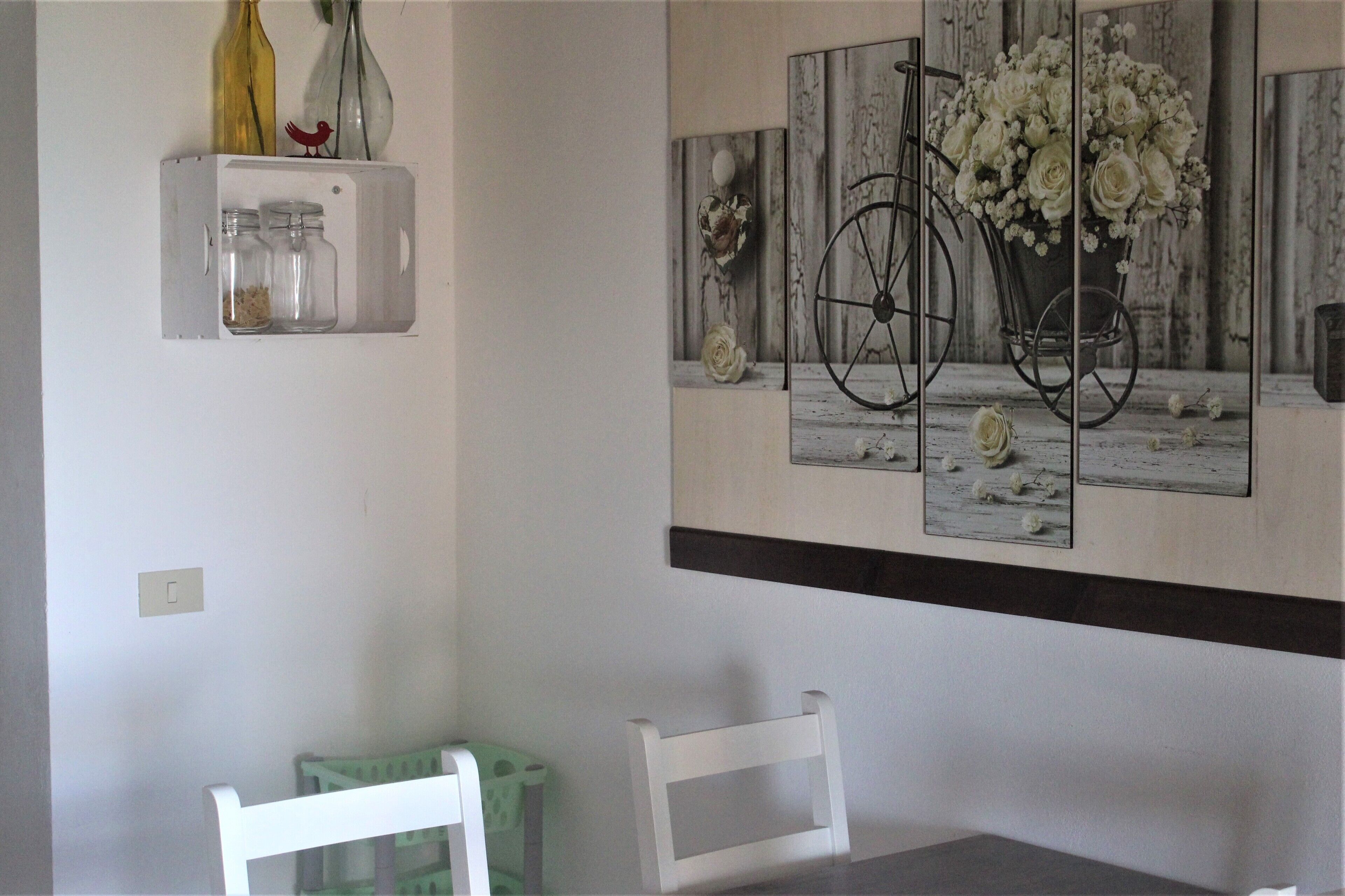 Un coin repas comprenant une table, des chaises, une étagère murale avec des bocaux et une œuvre d’art encadrée représentant un vélo et des fleurs.