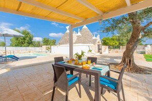 Un espace extérieur aménagé pour les repas, avec une table dressée pour deux personnes, une piscine et un bâtiment en pierre en arrière-plan.