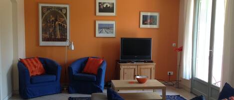 A living room with two blue armchairs, a sofa, a flat-screen TV on a wooden cabinet, and a window with curtains.