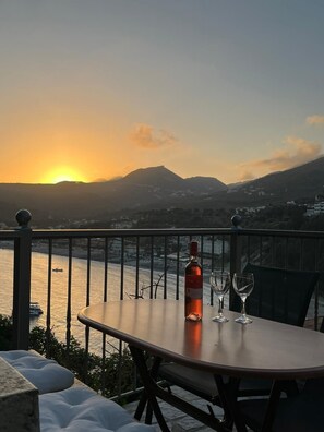 A table with a bottle and glasses, overlooking a body of water and mountains during sunset. - Paraskevi's Luxury Studios (Parga)