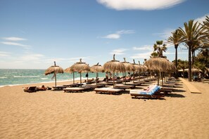 A beach with sun loungers and thatched umbrellas, palm trees, and a clear sky. - Amazing penthouse apartment within 3 minutes walk of Puerto Banus. (Marbella)