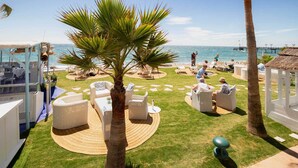 A beachside outdoor seating area with round tables and chairs, palm trees, and a view of the ocean.
