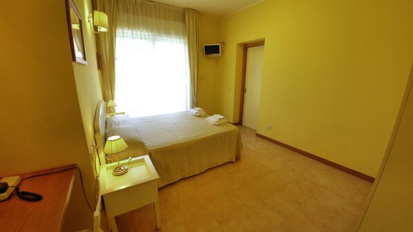 A small, bright room with a bed, bedside table, lamp, and a TV on the wall.