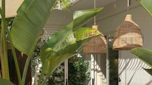 Un espacio de comedor al aire libre con sillas y mesas de madera, rodeado de plantas verdes grandes y lámparas de mimbre colgantes.