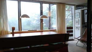 Un comedor con mesa de madera y bancos, ventanas amplias con cortinas y vista a árboles.