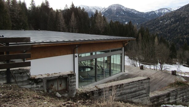 Un edificio moderno con techo inclinado y amplios ventanales de vidrio, rodeado por un bosque y montañas.