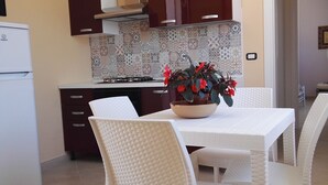 A kitchen with a white refrigerator, dark cabinets, a tiled backsplash, and a dining table with white chairs.