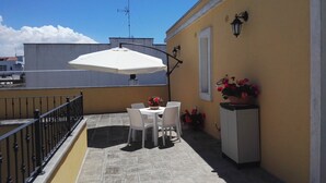 A balcony with a white umbrella, two white chairs, a small table, and a potted plant with red flowers.