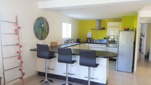 A modern kitchen with a central island, bar stools, a refrigerator, and a decorative wall clock. - Villa 8 people, swimming pool. Open view. Beaches 15 mins away, quiet. (Roquefort-Les-Pins)