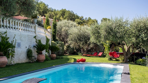 A swimming pool with a curved edge, surrounded by a well-maintained garden with various plants and a white wall with decorative tiles. - Mas de Provence en Riviera (La Roquette-sur-Var)