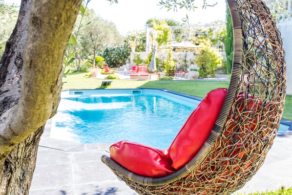A wicker chair with a red cushion by a swimming pool, surrounded by greenery and outdoor furniture. - Mas de Provence en Riviera (La Roquette-sur-Var)