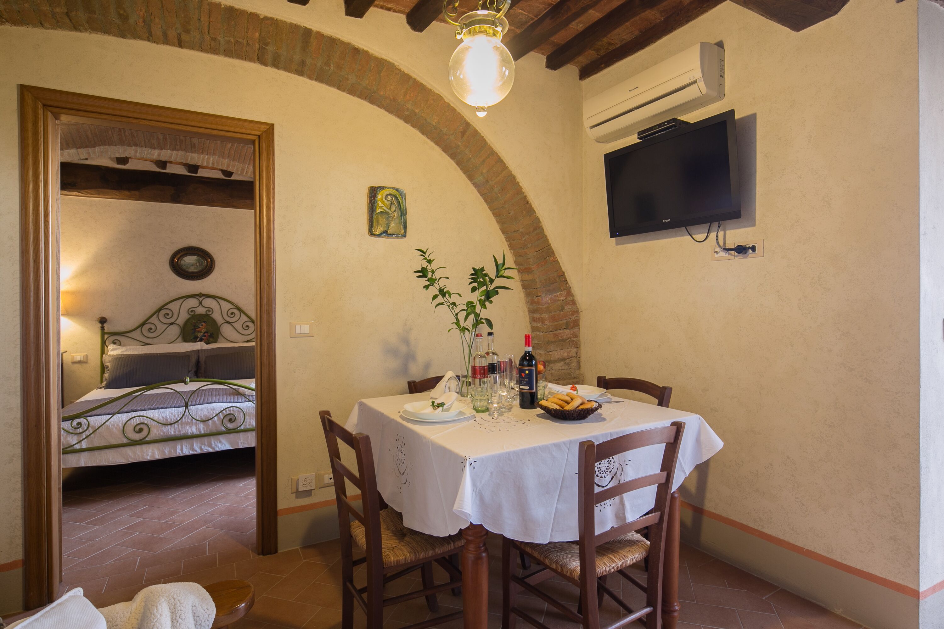 Un coin repas avec une table dressée pour deux personnes, un téléviseur fixé au mur et un lit visible à travers une arche.