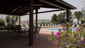 Zona de piscina al aire libre con sillas de descanso y mesas, rodeada de vegetación y colinas al fondo.