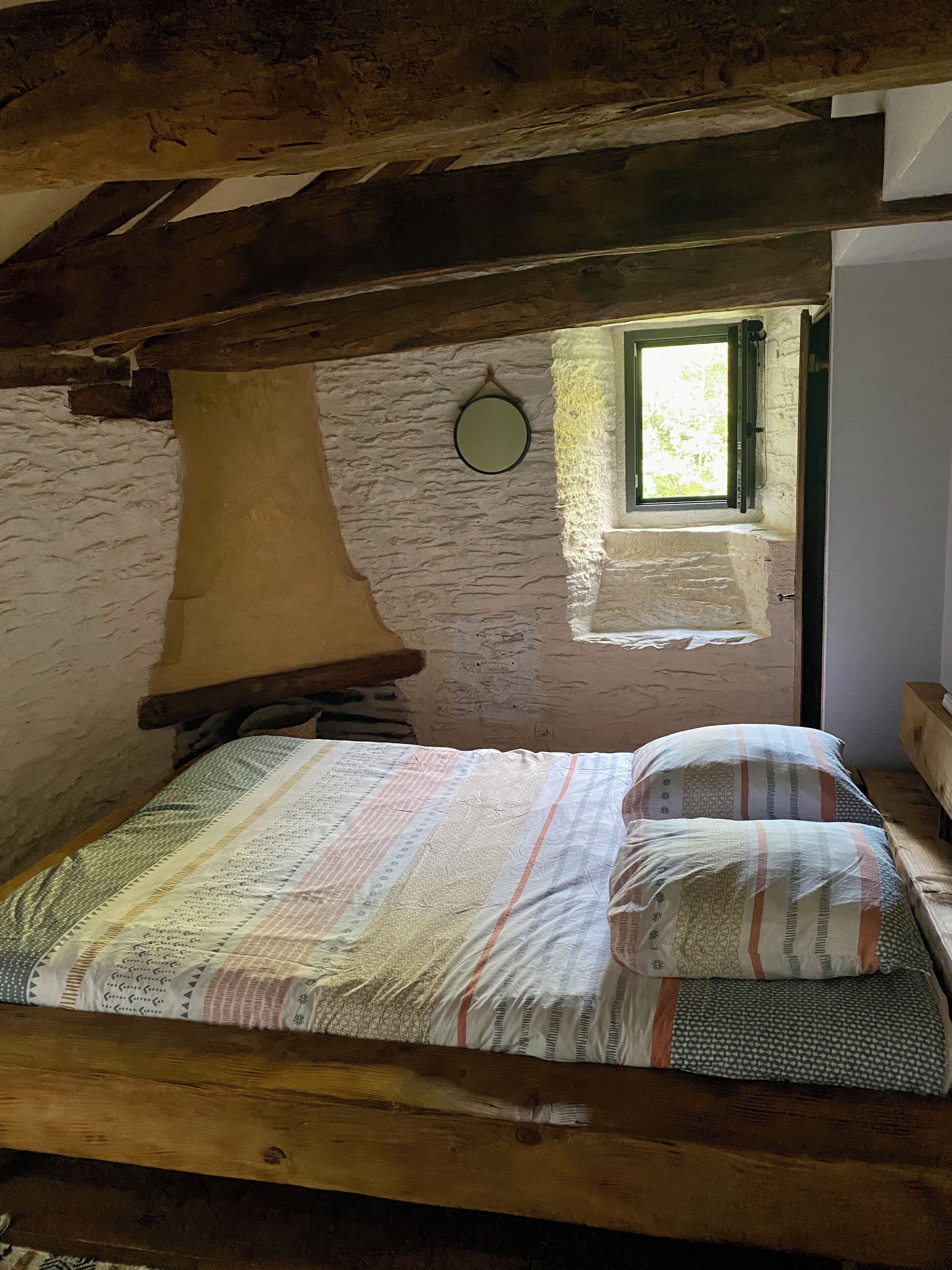 Una cama de madera rústica con una colcha y almohadas estampadas, una ventana pequeña con vistas a la vegetación y una pared de piedra.