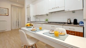 Une cuisine moderne avec des meubles blancs, un îlot central où sont disposés des aliments pour le petit-déjeuner, et une vue sur la salle à manger adjacente.