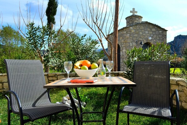 A table with a bowl of fruit and wine glasses, two wicker chairs, and a stone building in the background.