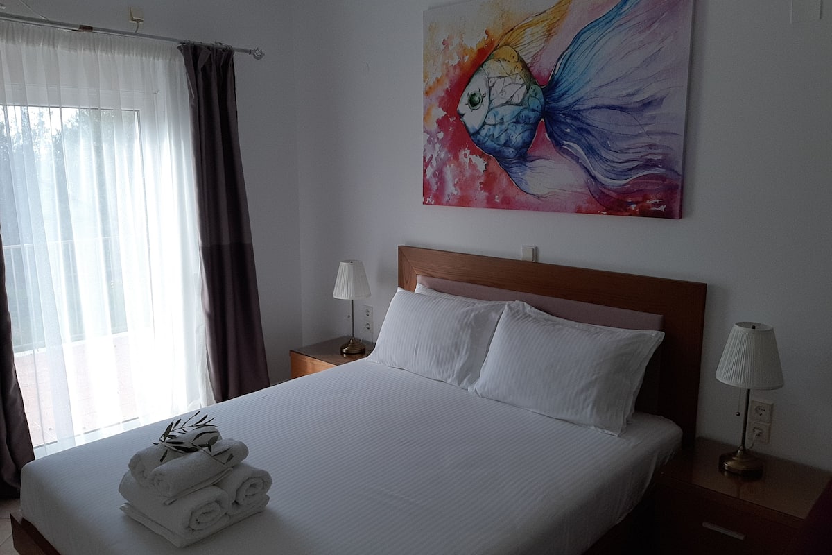 Ett hotellrum med en säng, två sängbord, ett fönster med gardiner och en färgstark tavla på väggen.
