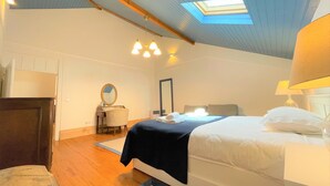 Un dormitorio con tragaluces, una cama con ropa de cama blanca, una mesita de noche con lámpara y una silla.