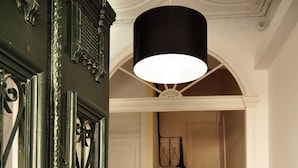 Eine Treppe mit hölzernen Stufen führt zu einer Zwischenebene, eine schwarze Metalttür mit aufwendigen Verzierungen und eine weiße Decke mit einer schwarzen Lampe.