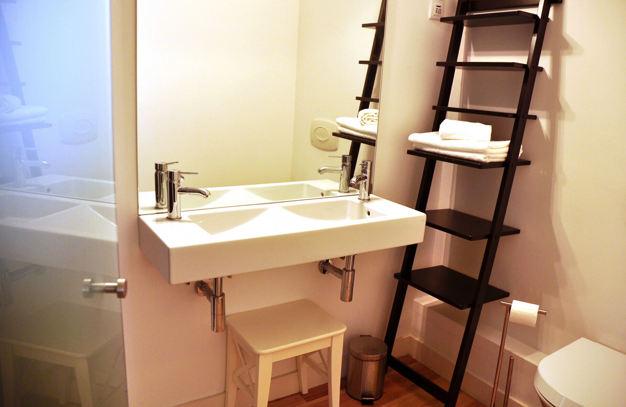 A bathroom with a white sink, a small stool, a black shelf, and a toilet.