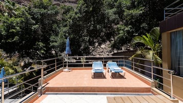 A rooftop terrace with two sun loungers, a blue umbrella, and a view of a rocky landscape.