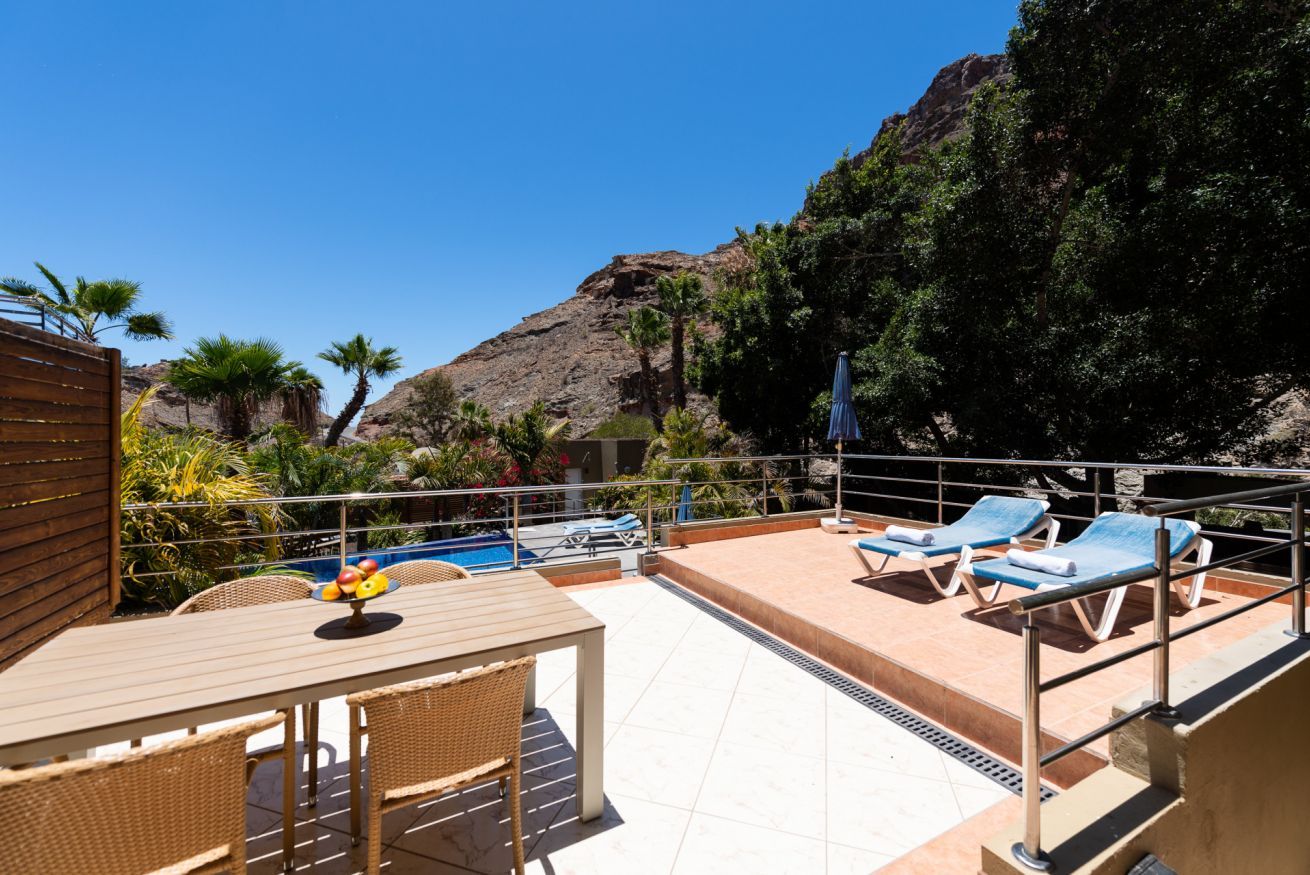 A rooftop pool area with lounge chairs, a table with fruit, and a view of a rocky hillside.
