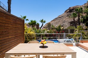 A wooden table with a fruit bowl, wicker chairs, a pool, and a mountain backdrop.