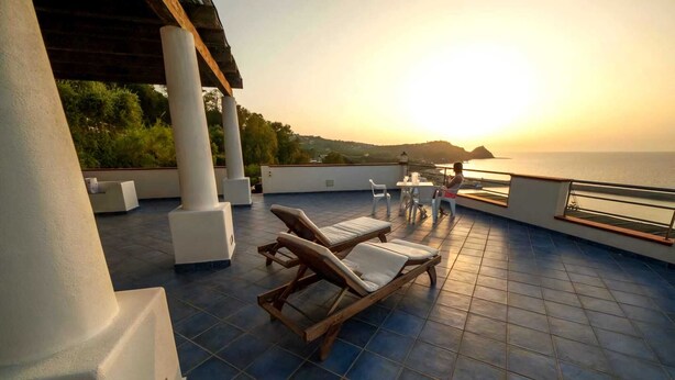 Una terraza en la azotea con dos sillones de madera, una mesa y vistas al mar al atardecer.