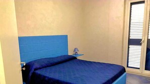 Un dormitorio con una cama azul, una mesita de noche con lámpara y una ventana con persianas.