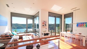 Uma sala de estar moderna com uma janela grande, uma área de jantar com cadeiras transparentes e piso de madeira.