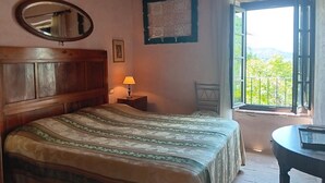 A bedroom with a wooden bed, a nightstand with a lamp, a mirror, a framed map on the wall, and a balcony with a view of greenery. - VILLA OLIVI. (Nocchi)