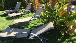 A row of sun loungers with metal frames and mesh seats, positioned on a well-maintained lawn with lush green grass and various tropical plants, including banana trees. - Semi-detached house in El Portil with private pool, free wifi, AC, BBQ  (El Portil)