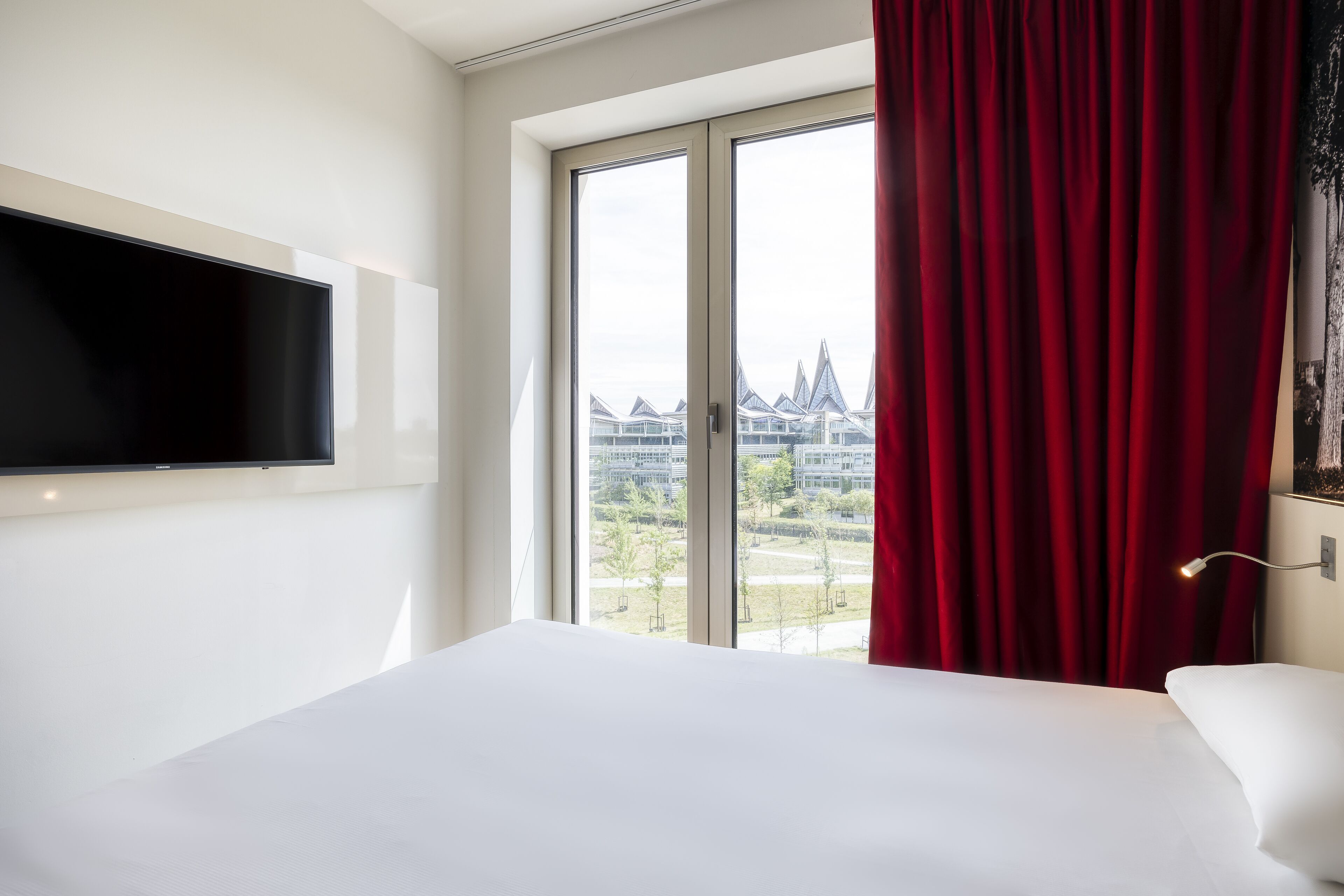 A hotel room with a large bed, a flat-screen TV mounted on the wall, and a window with red curtains.