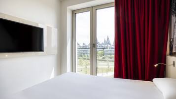 A hotel room with a large bed, a flat-screen TV mounted on the wall, and a window with red curtains.