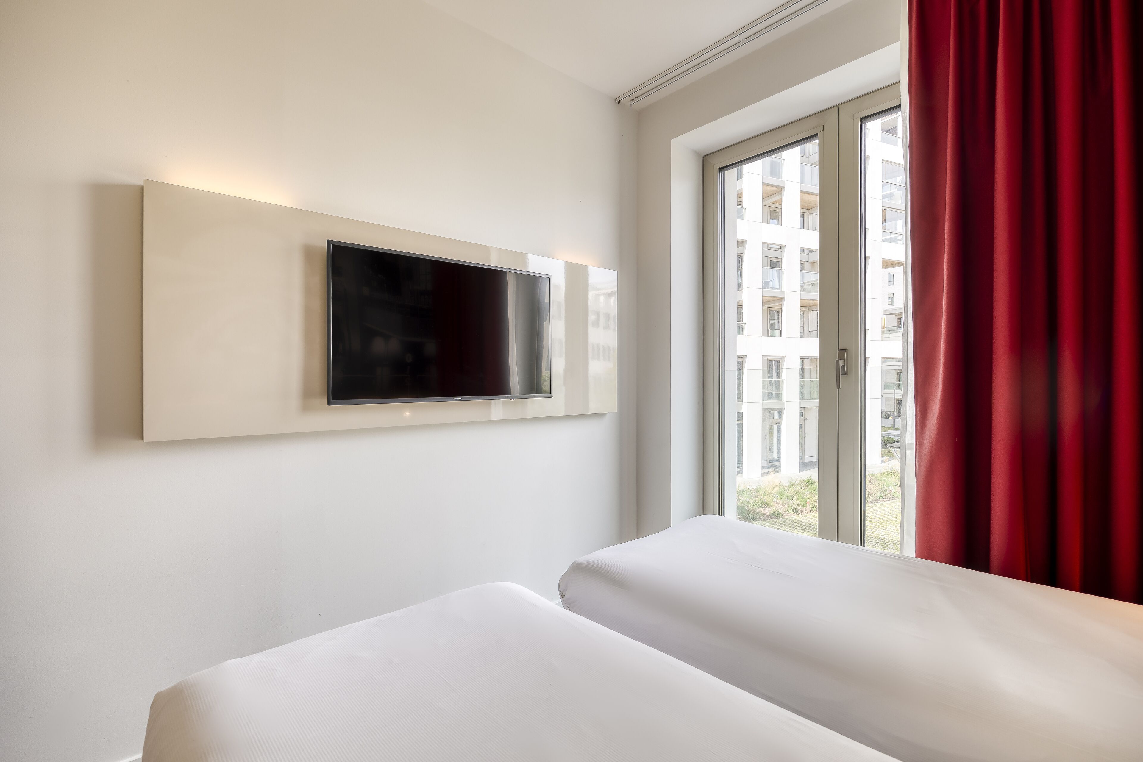 A hotel room with a bed, a mounted TV, and a window with red curtains.
