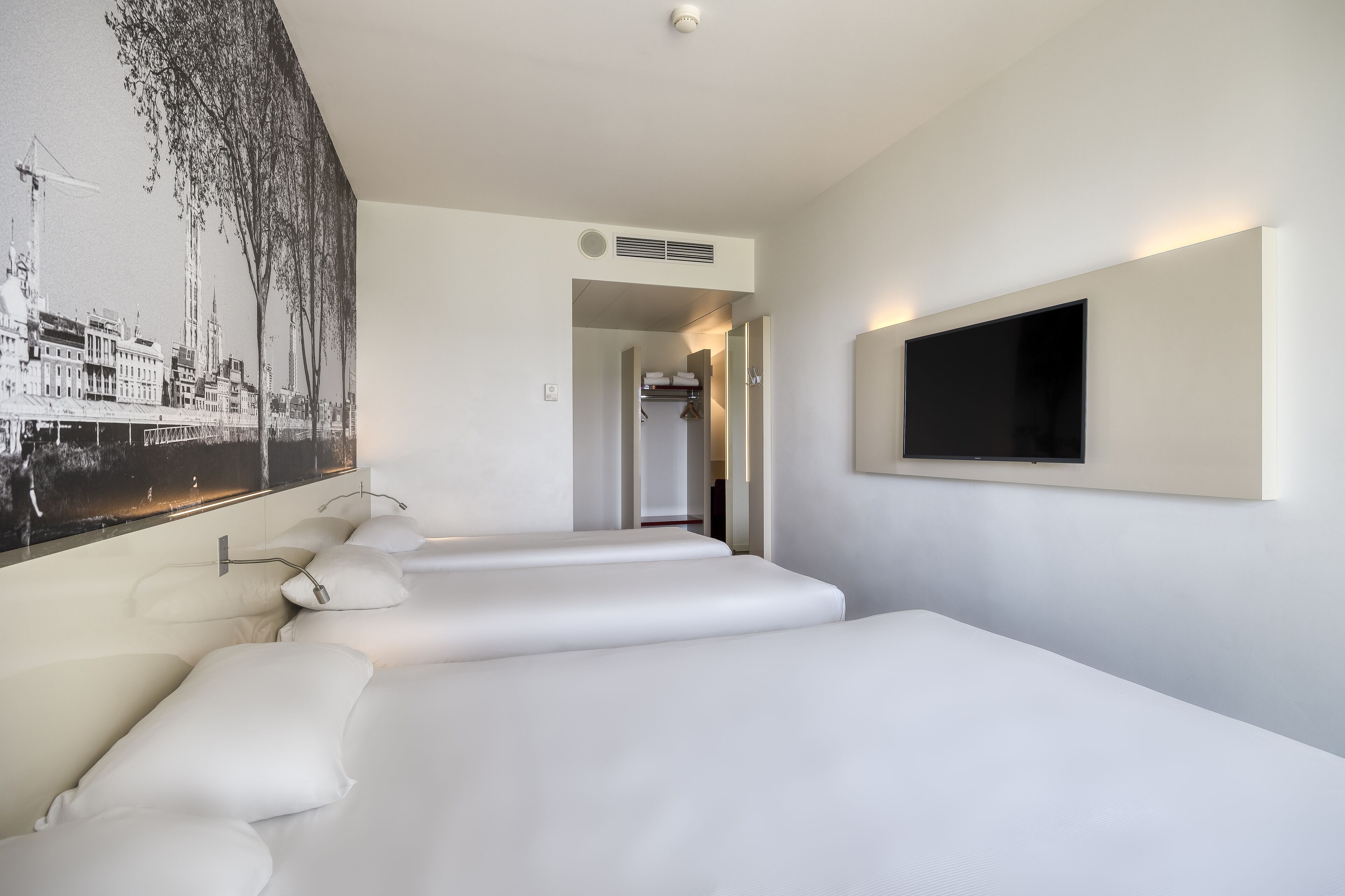 A hotel room with a large bed, a flat-screen TV mounted on the wall, and a cityscape mural on the wall.