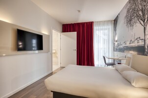 A modern hotel room with a large bed, a flat-screen TV mounted on the wall, a desk with a chair, and a large window with red curtains.