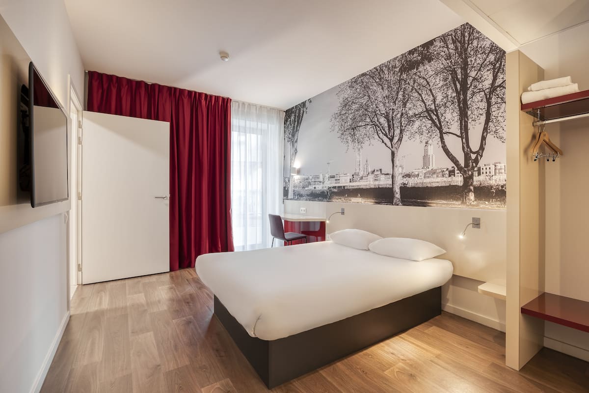 A modern hotel room with a large bed, a desk, and a cityscape mural on the wall.